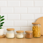 Bancada de cozinha pequena com potes de vidro e tampas de madeira, ao lado de planta decorativa e tábua de corte, em fundo de azulejos brancos.